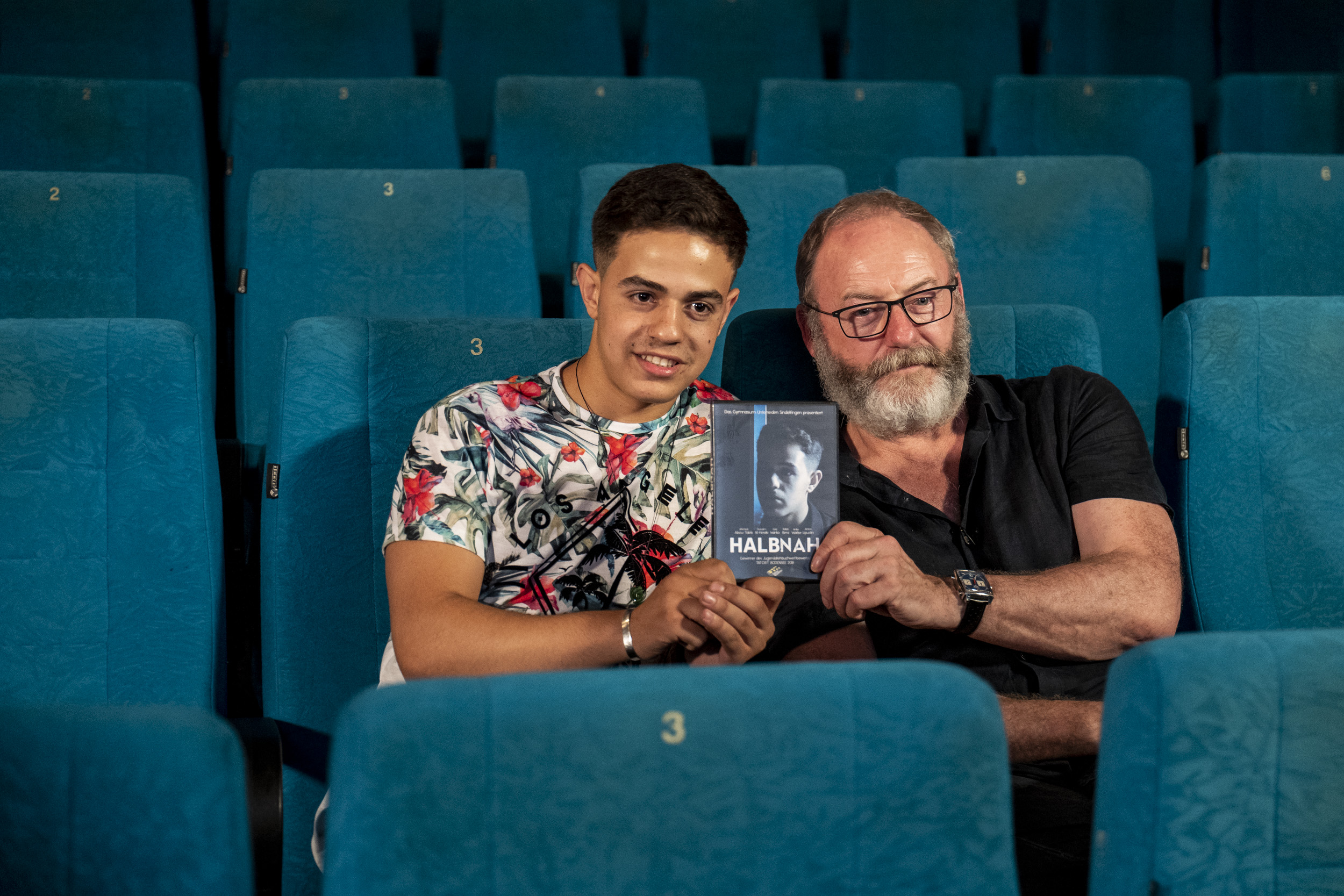 Liam Cunningham präsentiert ein Heft im Kinosaal zusammen mit einem Gast